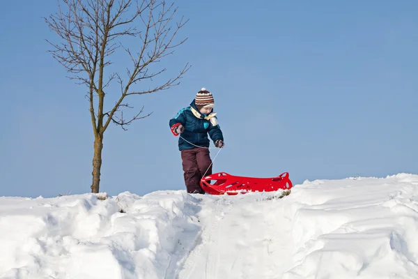 Menino puxando trenó de plástico vermelho para uma colina nevada — Fotografia de Stock