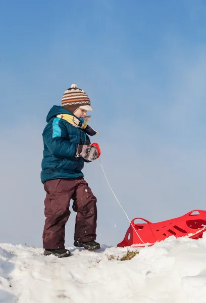 Jongen rood plastic slee trekken tot een besneeuwde heuvel — Stockfoto