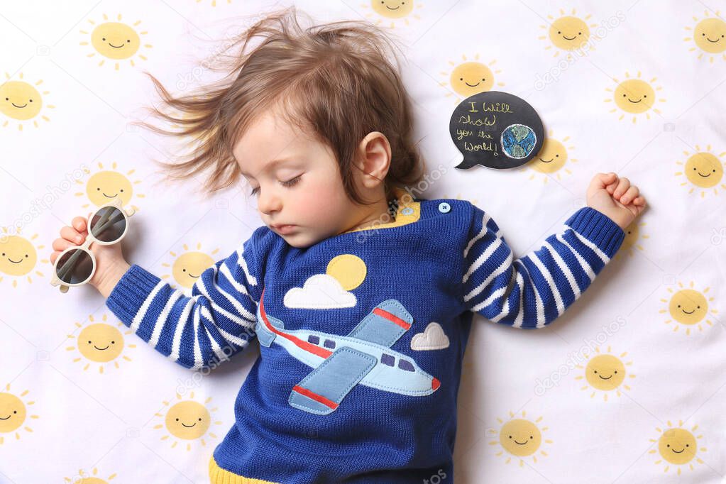 A sleeping baby wearing an airplane shirt and holding sunglasses dreamining of traveling, with a speech bubble saying I will show you the world.