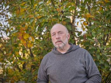 Arka planda elma ağacı olan, kendinden emin, ciddi ve kel bir adamın resmi.