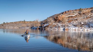 senior male wearing life jacket  is paddling expedition canoe in witer scenery of Horsetooth Reservoir in northern Colorado clipart
