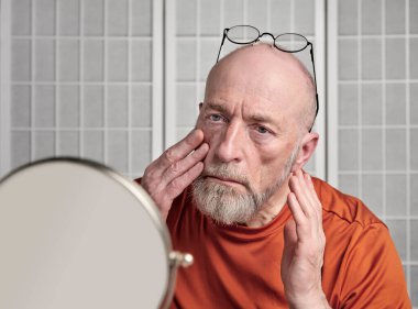 Kafası ve omuzları kel, sakallı yaşlı bir adamın portresi aynada yüzünü inceliyor, kendi kendine bakım kavramı.