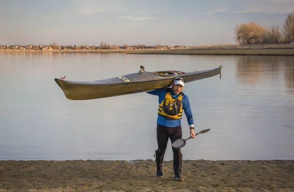Can yeleği giymiş orta yaşlı bir kürekçi uzun bir yarış kanosu ve bir göl sahilinde kanat küreği taşıyor.