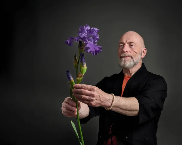 Head and shoulders portrait of bald and bearded senior man in kimono admiring or offering an iris flower, focus on flower
