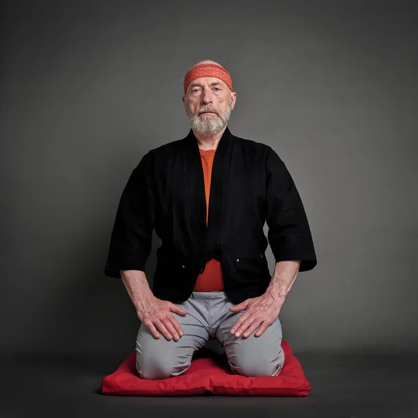 Head and shoulders portrait of bald and bearded senior man wearing a short kimono and sitting in a traditional Japanese seiza position on a cushion pad, contemplating, meditating or praying concept