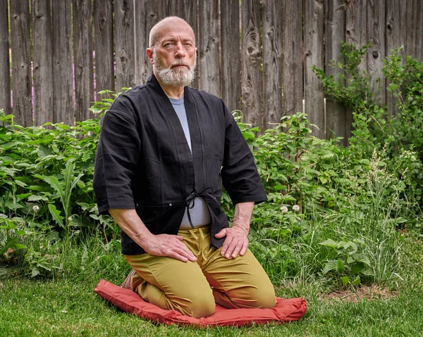 Head and shoulders portrait of bald and bearded senior man wearing a short kimono and sitting in a traditional Japanese seiza position on a cushion pad in a backyard, contemplating, meditating or praying concept