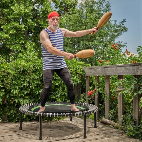 senior man is exercising on a mini trampoline with wooden Indian clubs in a backyard patio, fitness and rebounding concept