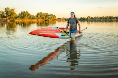 senior male paddler with a long and narrow racing stand up paddleboard on a calm lake at sunset.
