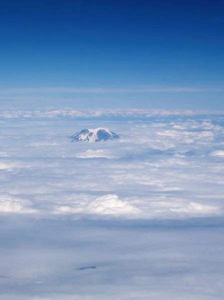 青い空と白い雲の層を通して雪の目で山頂の小さなビット — ストック写真