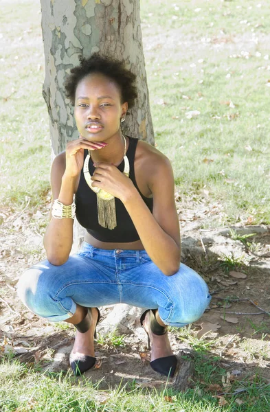 瘦小迷人的黑人少女蹲在牛仔裤里 站在树前的户外 — 图库照片