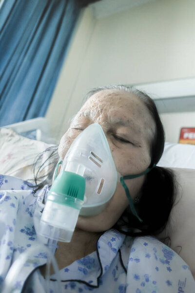 Asian female senior patient hospitalized and undergoing respiratory treatment using nebulizer in hospital bed