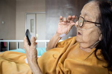 Southeast Asian female senior patient lying on hospital bed using smartphone for entertainment