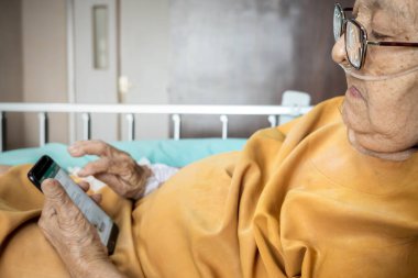 Southeast Asian female senior patient lying on hospital bed using smartphone for entertainment