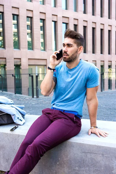 Genç sakallı adam cep telefonuyla konuşurken sokakta oturuyor.