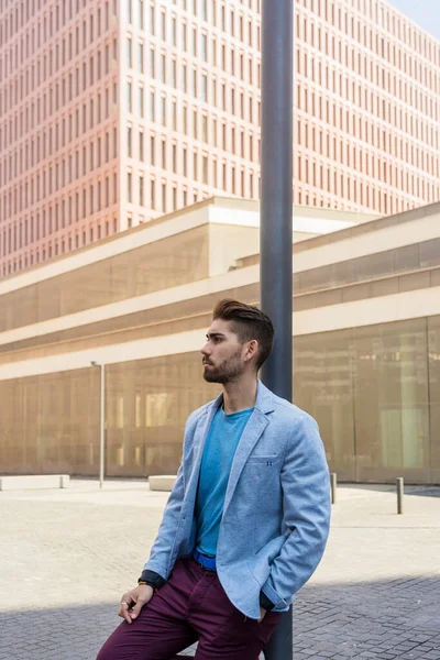 Bearded man, model of fashion, in urban background wearing casual clothes while leaning on a streetlight pole
