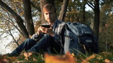 Sonbahar parkında ağacın altında düşen yaprakların üzerinde oturup tablet kullanan yakışıklı sakallı adam. Günbatımında güz mevsiminde sırt çantalı beyaz bir adam..