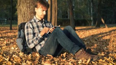 Sonbahar parkında ağacın altında düşen yaprakların üzerinde oturup tablet kullanan yakışıklı sakallı adam. Günbatımında sonbahar mevsiminde beyaz adam.