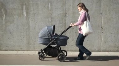 Kentte gri beton duvarın yanında bebek arabası ve bez bir alışveriş çantasıyla gezen aktif genç bir anne. Bakıcı anne bebek arabasında yeni doğan bebeğe bakıyor..
