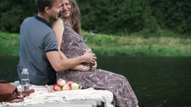 Hamile kadın ve kocası göldeki ahşap iskelede birlikte oturuyor, sarılıyor, öpüşüyor ve gülüyorlar. Genç bir aileden bebek beklerken çekilmiş gizli bir fotoğraf. Mutlu çift yaz pikniğinde açık havada.