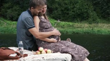 Hamile kadın ve kocası göldeki ahşap iskelede birlikte oturuyorlar. Genç bir aileden bebek beklerken çekilmiş gizli bir fotoğraf. Mutlu çift yaz pikniğinde açık havada.