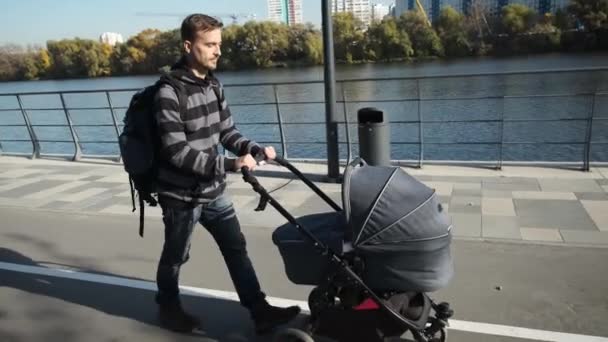 街の堤防を歩く新生児ベビーカーでハンサムな笑顔の父親の手持ち撮影 妊娠中の男休暇とともにPram — ストック動画