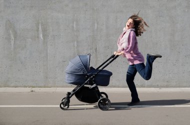 Yeni doğmuş bebek arabasını gri beton duvardan iterken dans eden heyecanlı genç kadın. Kendinden geçmiş anne şehirde bebek arabasıyla gezerken eğleniyor..