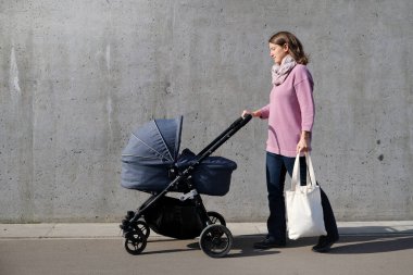Elinde bebek arabası ve tuvalle şehirde gri beton duvarın yanında yürüyen bir anne. Bakıcı anne bebek arabasında yeni doğan bebeğe bakıyor. Sıfır atık ebeveynlik.