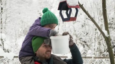 Feeding birds. Act of kindness. Cute child boy sits on dad shoulders and pouring sunflower seeds into feeder on tree at winter.
