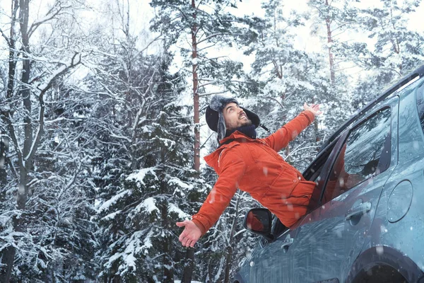 森の中で雪を楽しむ彼の車の窓の外に傾いてオレンジコートの幸せな楽観的な男のドライバー 寒い季節に田舎道で自動車を運転する 道路の旅 冬の休暇 自然を受け入れる — ストック写真