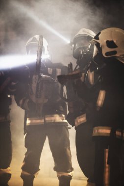 Profesyonel itfaiyeciler tam teçhizat, oksijen maskesi ve acil durum kurtarma araçları, dairesel hidrolik ve gaz testeresi, balta ve balyoz takıyorlar. Arka planda duman ve itfaiye araçları.