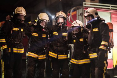 Profesyonel itfaiyecilerden oluşan bir grup. Üniforma giyen itfaiyeciler, koruyucu kasklar ve oksijen maskeleri takıyorlar. Arka planda duman ve itfaiye araçları.