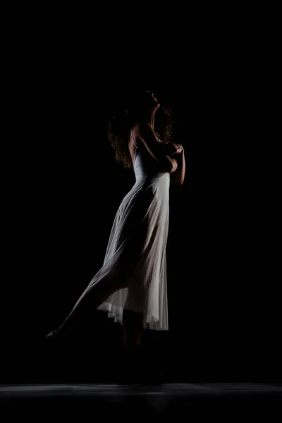 巻き毛のバレエポーズの女の子 サイドライトシルエットのバレリーナで白いドレスと黒のブーツに黒の背景 — ストック写真