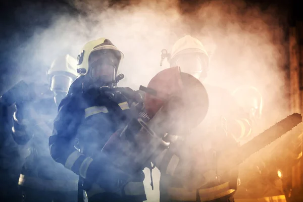 Group of professional firefighters wearing full equipment, oxygen masks, and emergency rescue tools, circular hydraulic and gas saw, axe, and sledge hammer. smoke and fire trucks in the background.