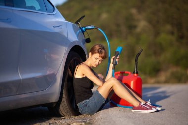 Üzgün kız yerde elektrikli arabanın yanında oturuyor. Şarj kablosu ve kırmızı gaz tüpü duruyor..