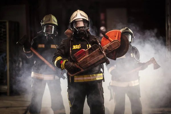 Group of professional firefighters wearing full equipment, oxygen masks, and emergency rescue tools, circular hydraulic and gas saw, axe, and sledge hammer. smoke and fire trucks in the background.