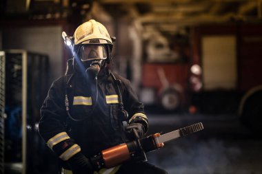 Arka planda tam teçhizat, oksijen maskesi ve hidrolik kesici alet, duman ve itfaiye araçları olan itfaiyeci portresi var..