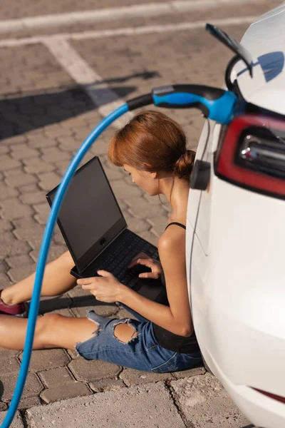 Lány Földön Vár Miközben Elektromos Autója Töltődik Laptopon Dolgozni — Stock Fotó