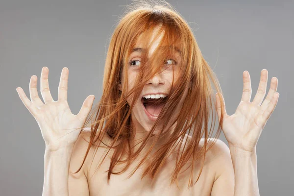 Güzel, koyu turuncu, rüzgarlı saçlı kız. Şaşırmış yüz ifadesi ve gri arka planda el hareketi ile stüdyo portresi.