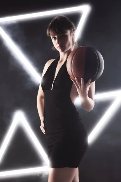 穿着漂亮的黑色连衣裙 拿着篮球的漂亮姑娘 带有白色霓虹灯的雾背景下的工作室肖像 — 图库照片