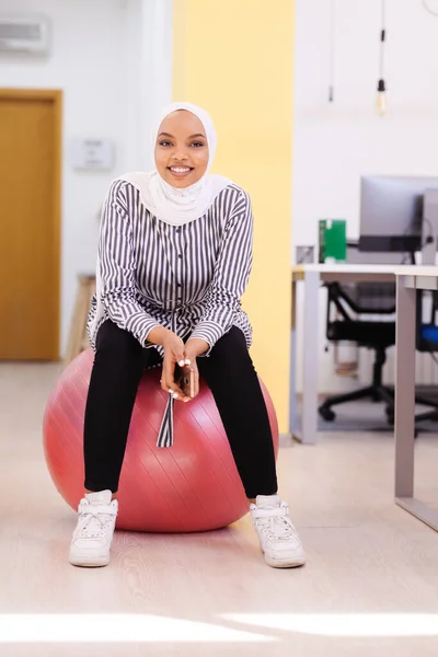 African american muslim girl with hijab sitting on balance ball n a modern office.