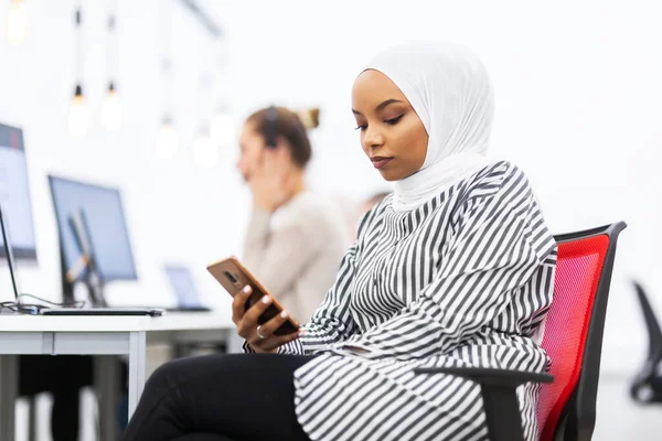 African american muslim girl with hijab working on a phone in modern office. Caucasian girl colleague with headset in the background
