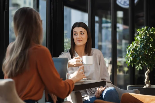 Two young business women in a cafe having one on one meeting. Friends after work talking gossiping and having coffee at a window table on a sunny day.