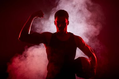 Siluet basketbol oyuncusu tarafı kırmızı renkli, arka planda duman olan bir topu tutuyor. Ciddi Afrikalı Amerikalı adam kaslarını gösteriyor.