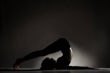 Fit balerin kız yalan söylüyor ve yoga pozları veriyor. Koyu arkaplana karşı yan aydınlatma silueti.