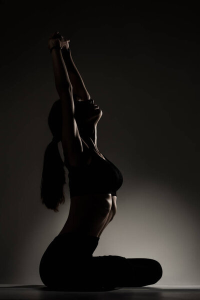 Fit girl resting and stretching after practicing yoga poses. Side lit half silhouette against dark background.