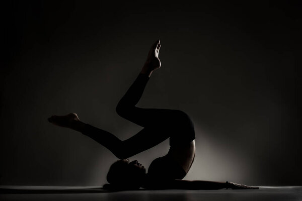 Fit ballerina girl lying and stretching practicing yoga poses. Side lit silhouette against dark background.