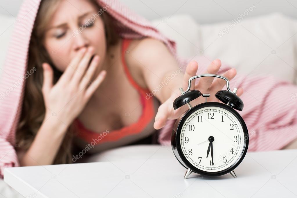 Sleepy young woman in bed with eyes closed extending hand to alarm clock at home
