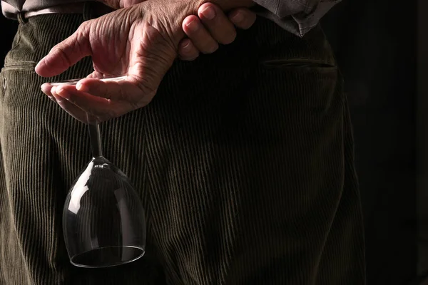 Closeup of a man wearing brown coduroy trousers and shirt with the sleeves rolled up holding a wine glass behind his back.