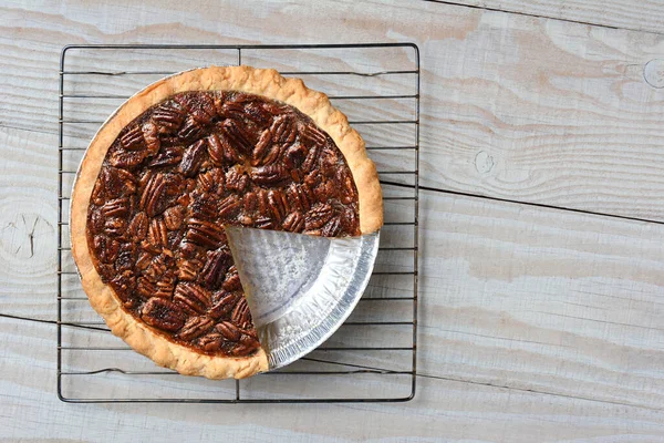 Overhead shot of a pecan pie with a slice cut out on a cooling rack. Horizontal format on a rustic whtie kitchen table.