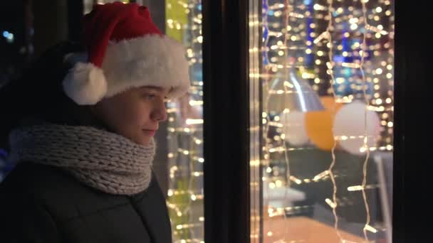 Хлопчик в капелюсі Сантас дивиться у вікно магазину — стокове відео
